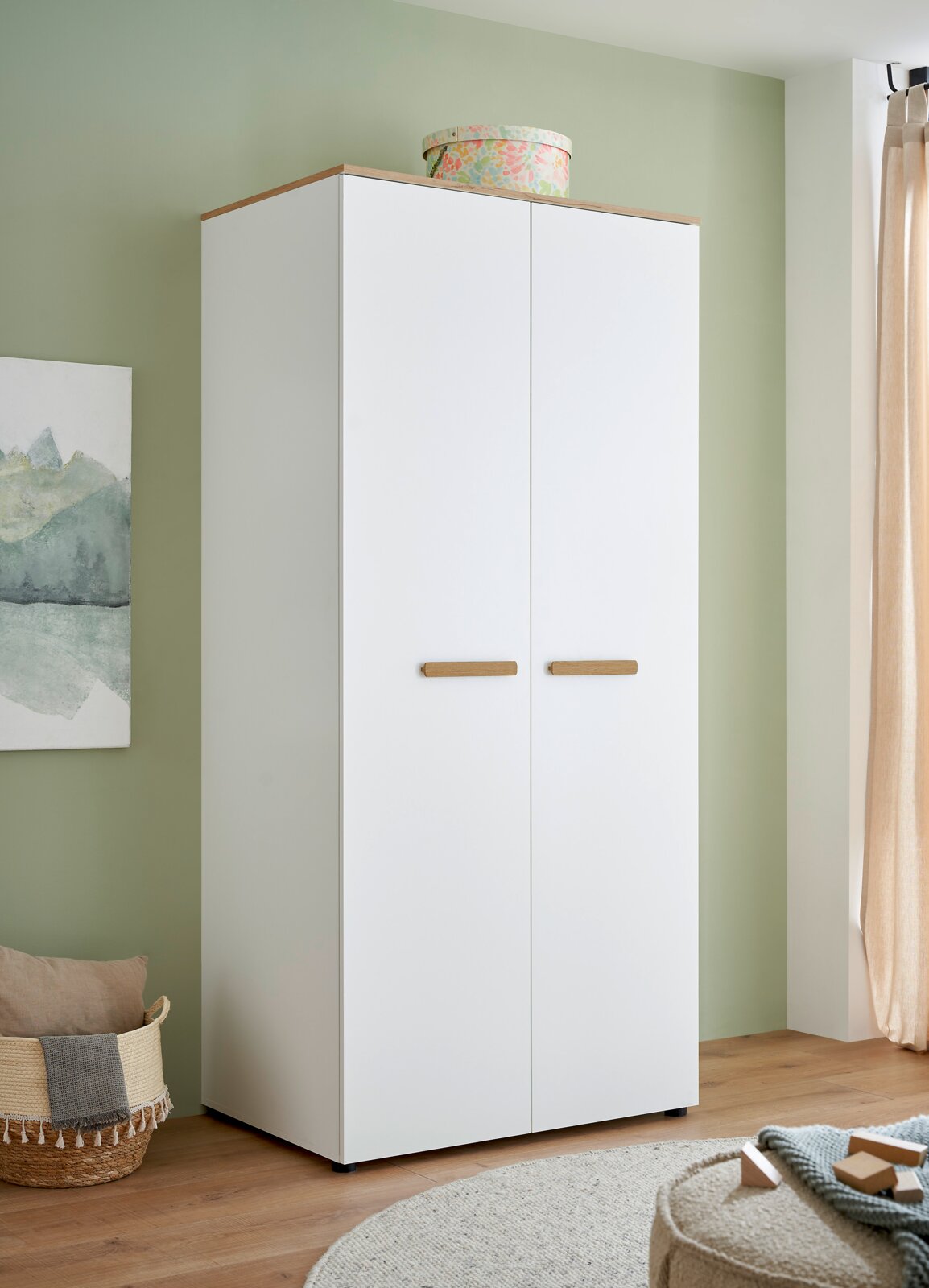 MyBaby Kleiderschrank ASTEA Weißer zweitüriger Kleiderschrank mit Holzoptik-Oberplatte und Holzgriffen, im Zimmer vor grüner Wand – frontale Ansicht mit leichter Perspektive von links.