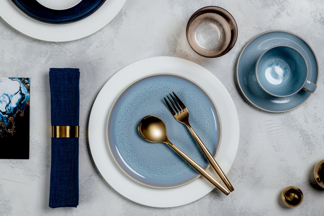 Elegantes Tischarrangement mit blauem Geschirr, goldfarbenem Besteck und einem Wasserglas aus der Vogelperspektive.