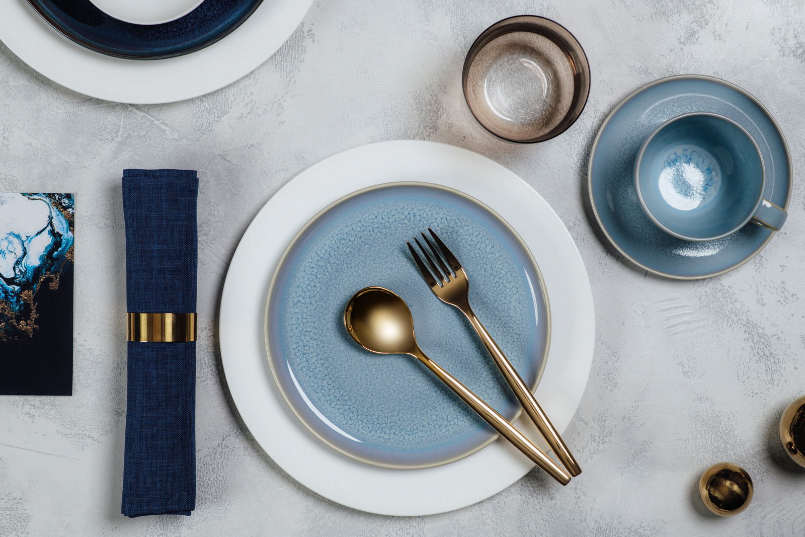 Elegantes Tischarrangement mit blauem Geschirr, goldfarbenem Besteck und einem Wasserglas aus der Vogelperspektive.