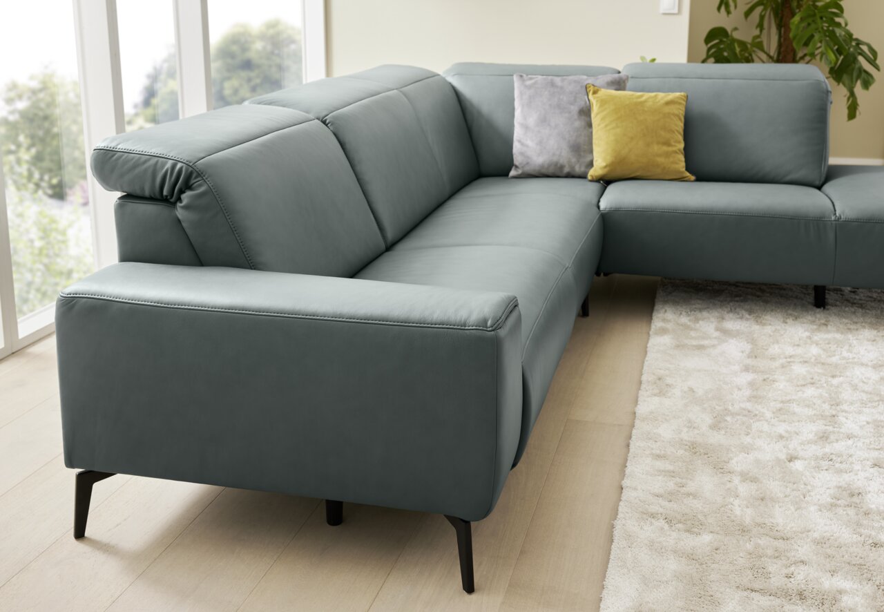Ecksofa Musterring MR 1320 in grauem Leder, seitliche Perspektive, mit zwei Kissen in Gelb und Grau auf hellem Holzboden und Teppich.