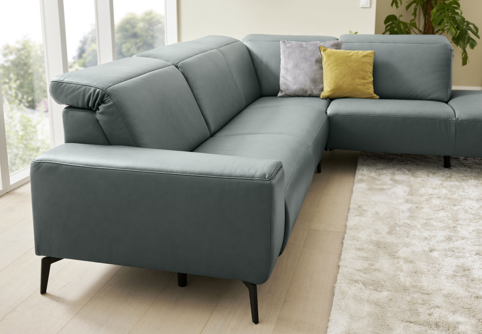 Ecksofa Musterring MR 1320 in grauem Leder, seitliche Perspektive, mit zwei Kissen in Gelb und Grau auf hellem Holzboden und Teppich.