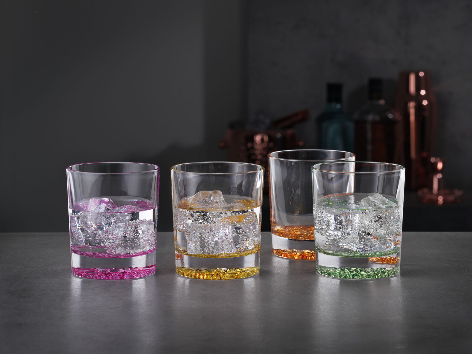 Vier bunte Trinkbecher mit Eiswürfeln, von vorne fotografiert, auf einer dunklen Oberfläche.