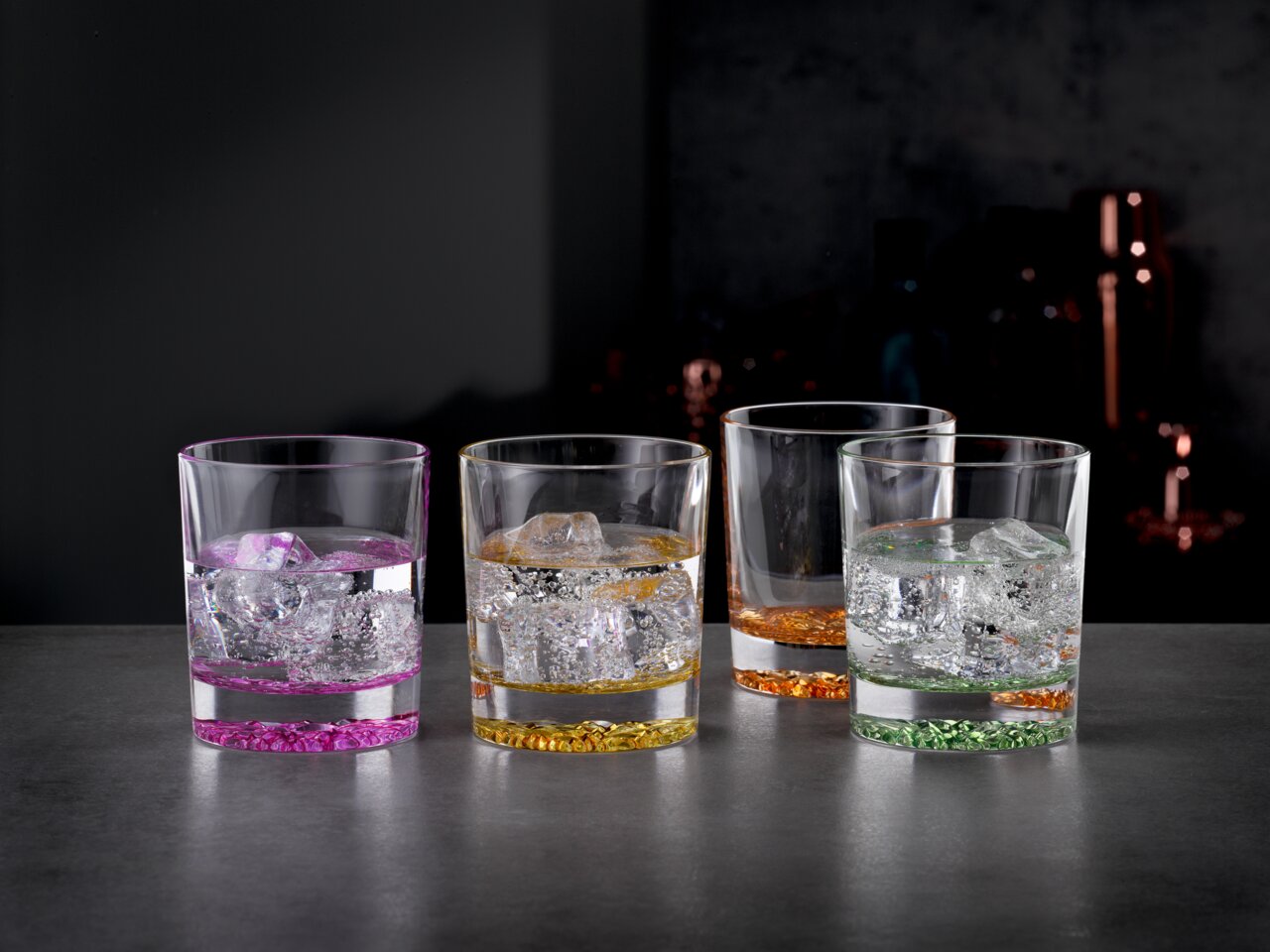 Vier bunte Trinkbecher mit Eiswürfeln, von vorne fotografiert, auf einer dunklen Oberfläche.