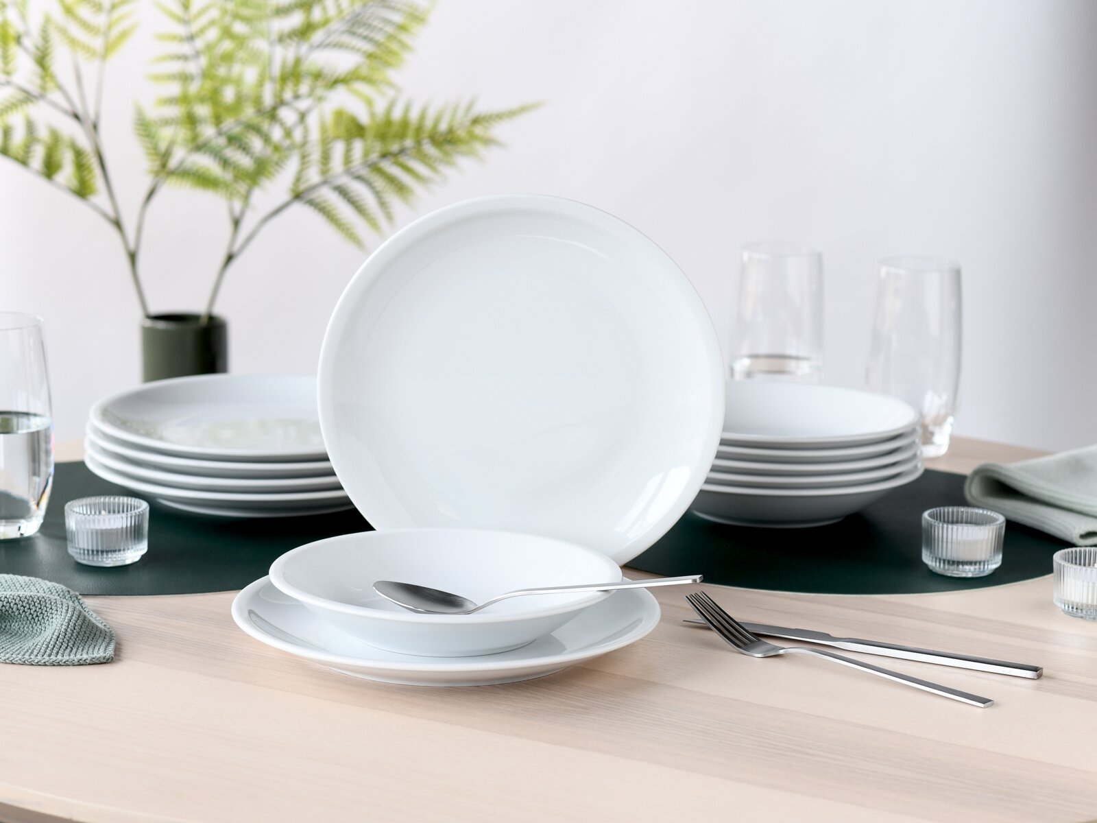 creaTable Tafelservice 12-tlg MILANO Weißes, minimalistisches Porzellan-Geschirrset mit Tellern und Schalen auf gedecktem Tisch, Frontansicht mit leicht erhöhter Perspektive; im Hintergrund Gläser, Besteck und grüne Pflanze.