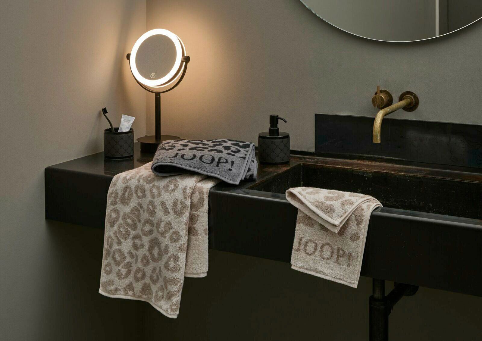 JOOP! Handtuch LEO Luxuriöses Badezimmer mit zwei JOOP! Handtüchern im Leo-Design, eines in Beige und eines in Grau, drapiert über einem modernen Waschbecken. Die Perspektive zeigt die Handtücher von vorne, mit einem beleuchteten Spiegel im Hintergrund.