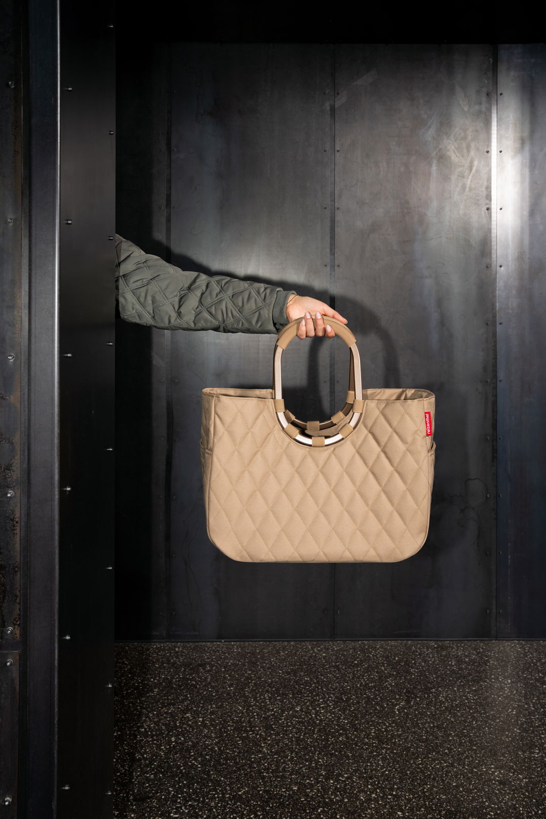 Beige Tasche mit gestepptem Muster, von einer Hand in einem grünen Ärmel gehalten, vor einem dunklen Hintergrund. Seitenansicht.