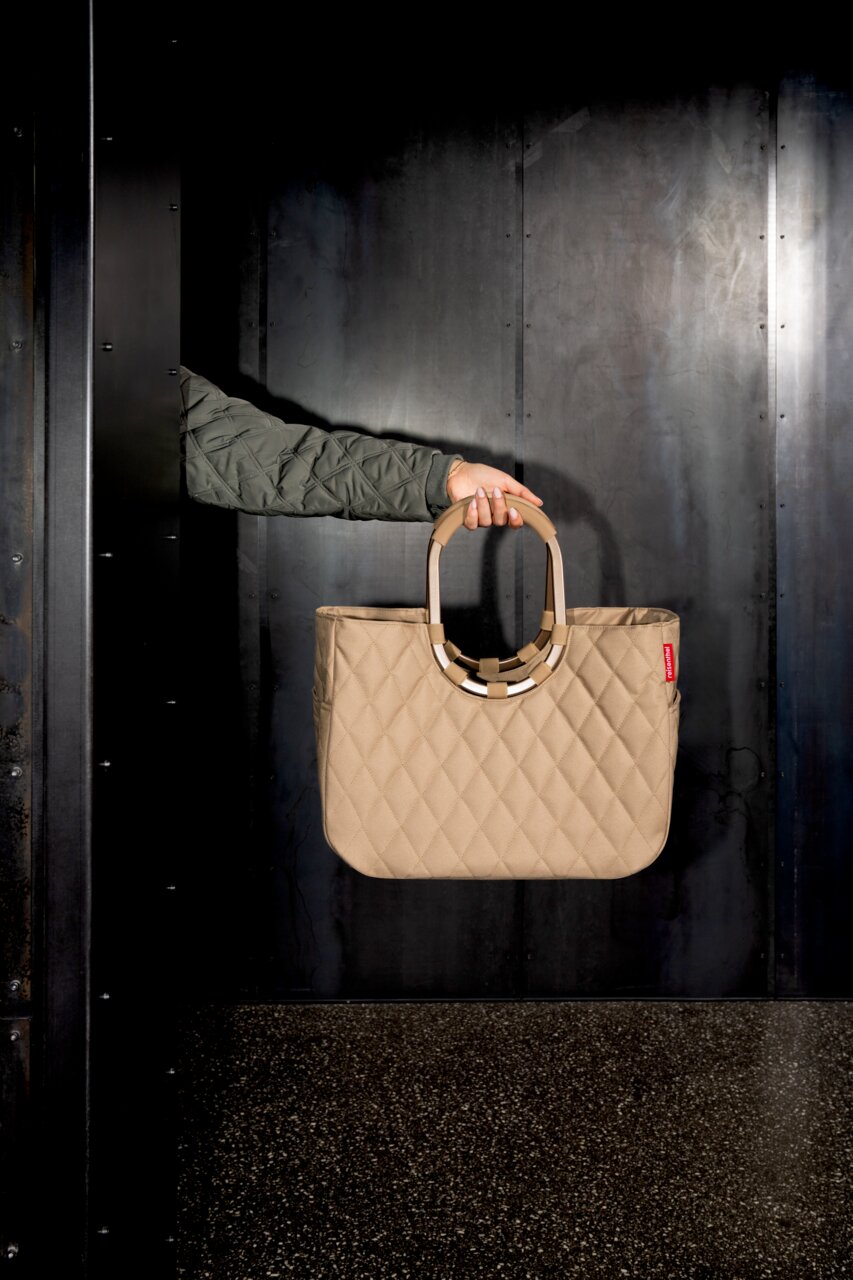 Beige Tasche mit gestepptem Muster, von einer Hand in einem grünen Ärmel gehalten, vor einem dunklen Hintergrund. Seitenansicht.