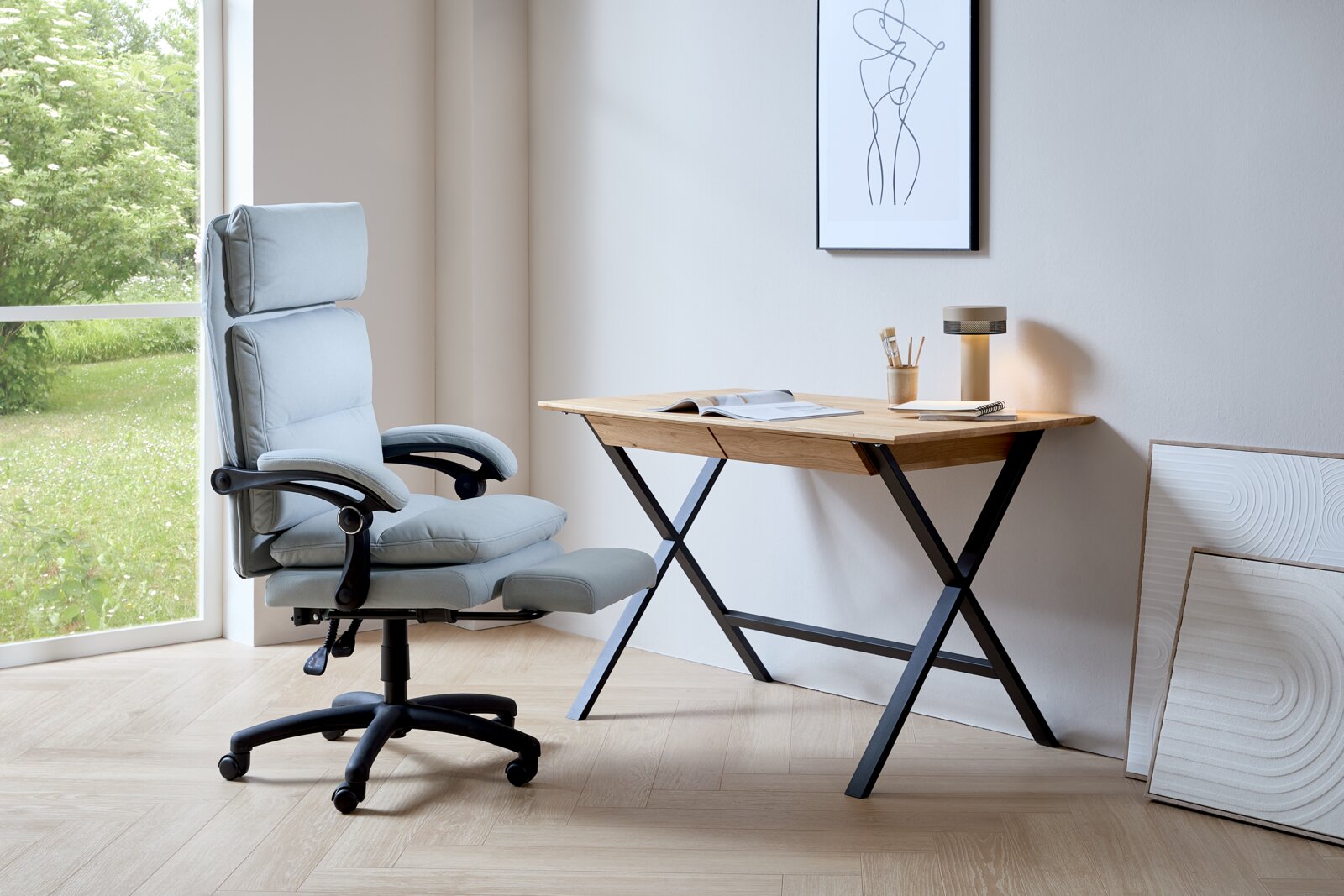 Modern bureau met houten blad en zwarte metalen poten, gezien vanaf de zijkant. Ernaast staat een ergonomische bureaustoel voor een groot raam met uitzicht op het groen.