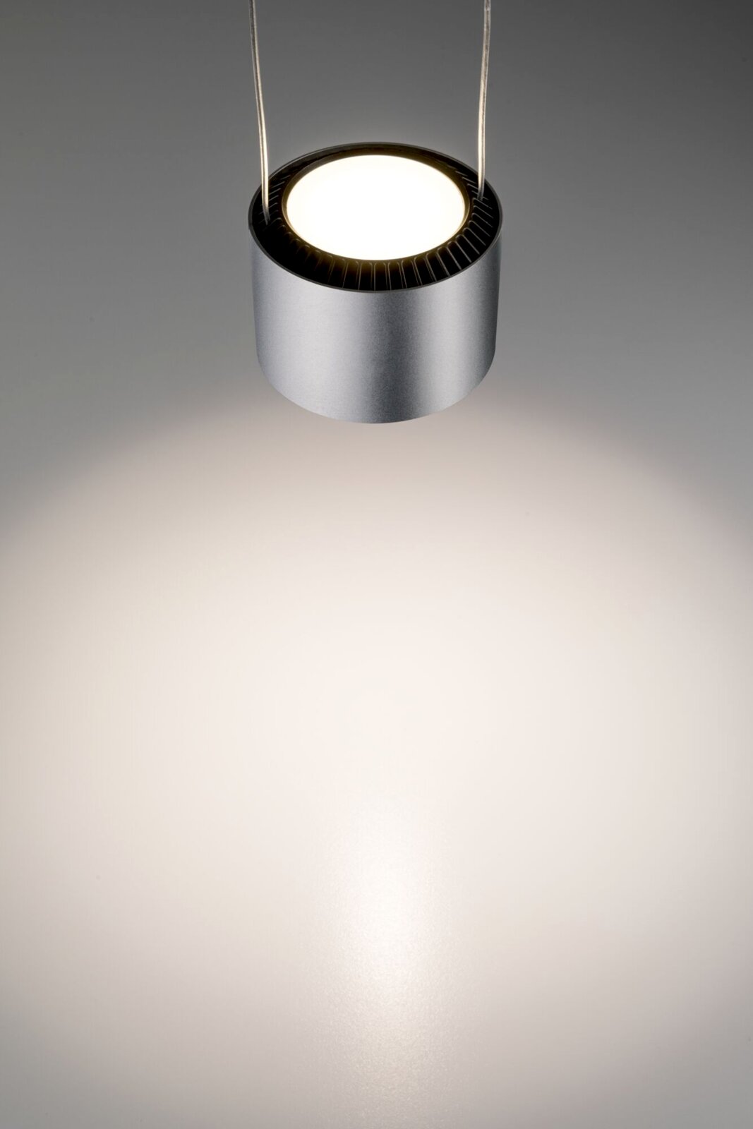 Moderne, zylindrische Pendelleuchte in Silber, von oben fotografiert, mit warmem Lichtstrahl auf einer grauen Oberfläche.