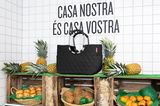 Schwarze, rautenförmig gesteppte Tasche, präsentiert auf einem Marktstand mit Ananas und Kokosnüssen, seitliche Perspektive.