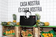 Schwarze, rautenförmig gesteppte Tasche, präsentiert auf einem Marktstand mit Ananas und Kokosnüssen, seitliche Perspektive.
