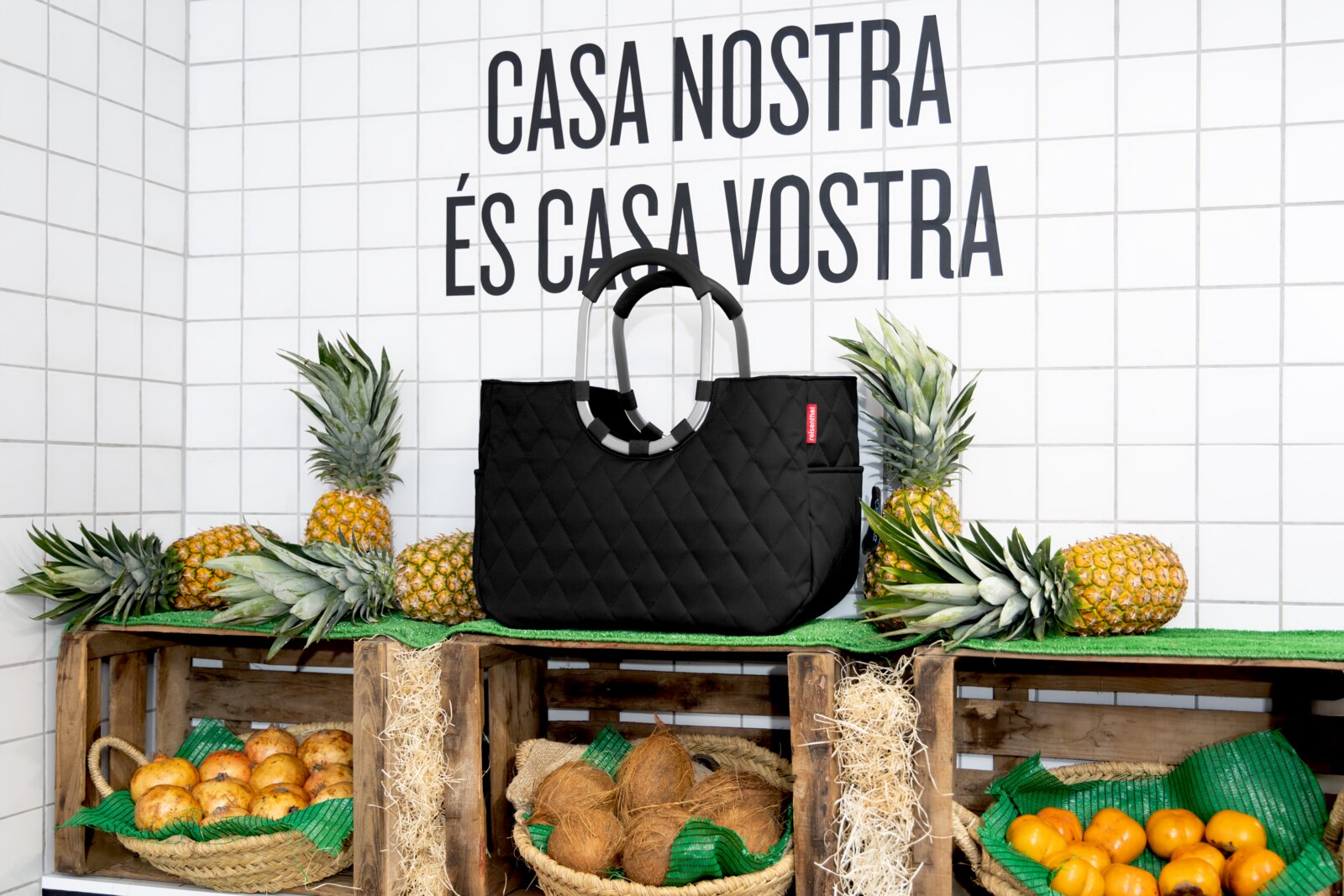 Schwarze, rautenförmig gesteppte Tasche, präsentiert auf einem Marktstand mit Ananas und Kokosnüssen, seitliche Perspektive.
