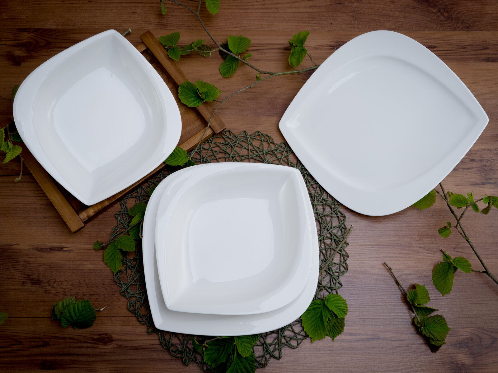 Dreiteiliges Tafelservice-Set aus weißem Porzellan, bestehend aus zwei quadratischen und einem ovalen Teller, auf einem Holztisch mit grünen Blättern dekoriert, aus der Vogelperspektive fotografiert.