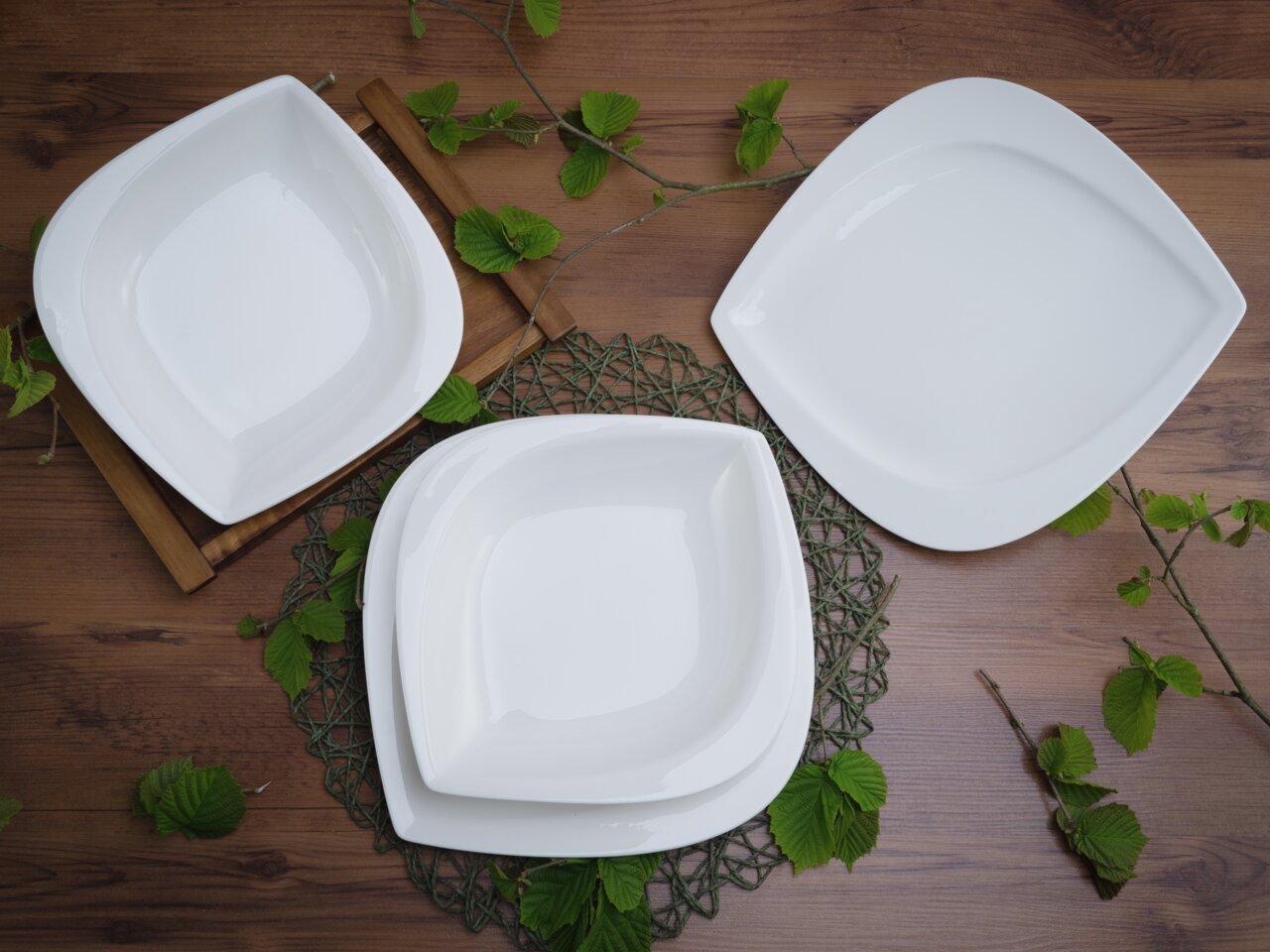 Dreiteiliges Tafelservice-Set aus weißem Porzellan, bestehend aus zwei quadratischen und einem ovalen Teller, auf einem Holztisch mit grünen Blättern dekoriert, aus der Vogelperspektive fotografiert.