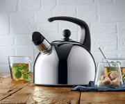 Silberner Flötenkessel mit schwarzem Griff und Pfeifaufsatz, auf einem Holztisch stehend, mit zwei Gläsern Tee im Vordergrund. Perspektive von vorne.