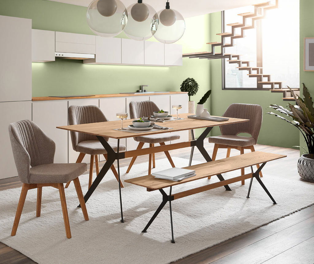Moderner Essbereich mit einem Esstisch und Sitzbank aus Holz und Metall, umgeben von gepolsterten Stühlen in einem hellen Raum mit grüner Wand und Treppe im Hintergrund. Perspektive: seitliche Ansicht.
