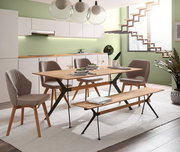 Moderner Essbereich mit einem Esstisch und Sitzbank aus Holz und Metall, umgeben von gepolsterten Stühlen in einem hellen Raum mit grüner Wand und Treppe im Hintergrund. Perspektive: seitliche Ansicht.