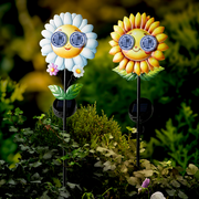 Zwei fröhliche Solar-Stabbeleuchten in Form von Sonnenblumen mit lächelnden Gesichtern und Sonnenbrillen, aufgestellt in einem Garten. Vorderansicht.