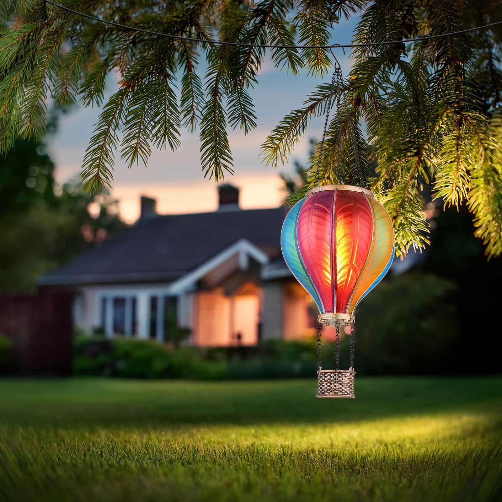 Ambia Garden Solarleuchte Bunte Solarleuchte in Form eines Heißluftballons, aufgehängt an einem Baumzweig, mit einem Haus im Hintergrund bei Dämmerung.