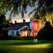 Bunte Solarleuchte in Form eines Heißluftballons, aufgehängt an einem Baumzweig, mit einem Haus im Hintergrund bei Dämmerung.