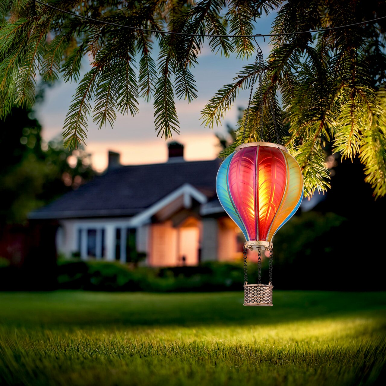 Bunte Solarleuchte in Form eines Heißluftballons, aufgehängt an einem Baumzweig, mit einem Haus im Hintergrund bei Dämmerung.