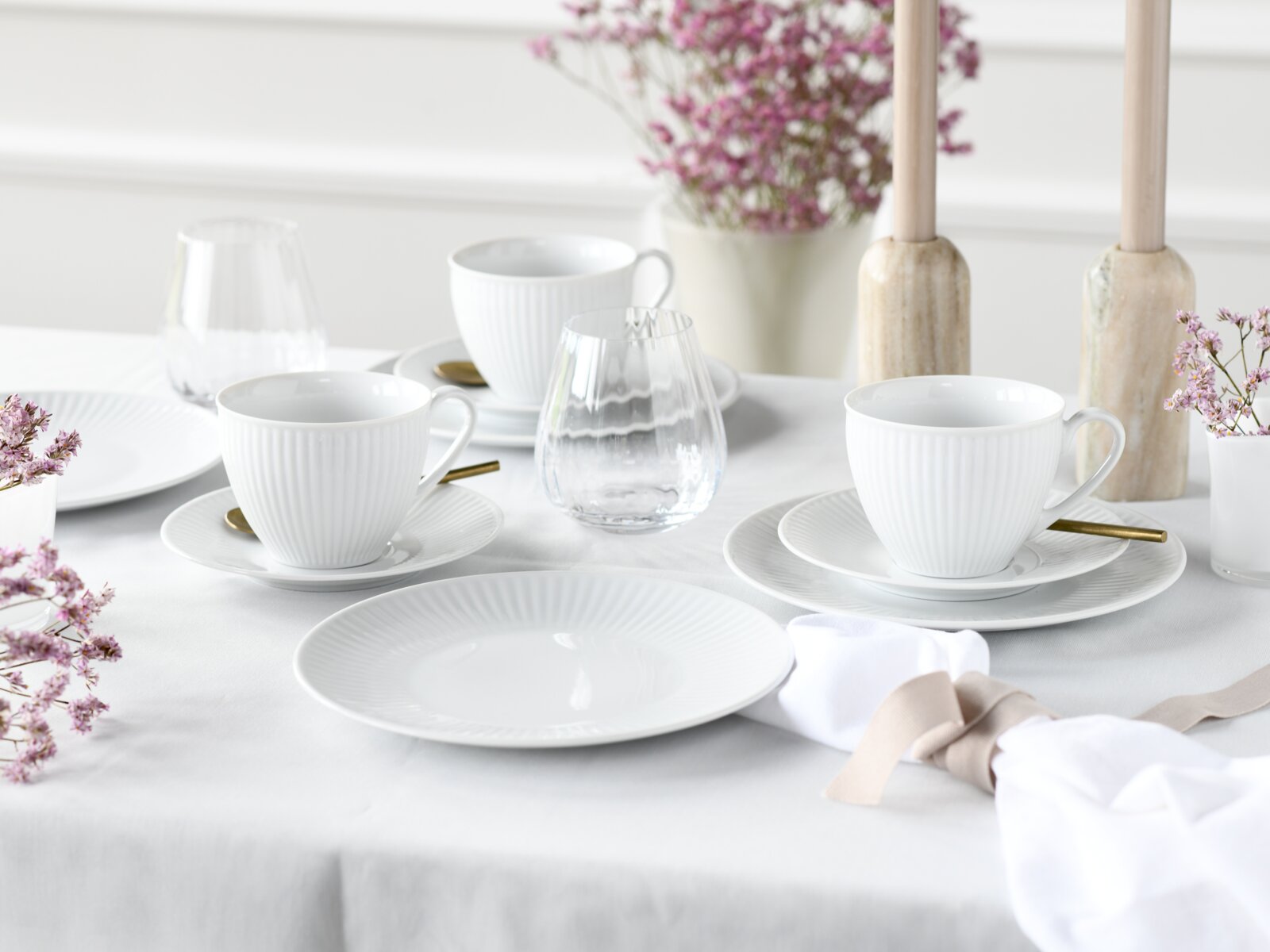 Schlicht gedeckter Tisch mit weißem geripptem Porzellan-Geschirr (Tassen mit Untertassen und Teller), klaren Wassergläsern und goldfarbenem Besteck, dekoriert mit rosa Blumen und Holzkerzenständern, fotografiert aus leicht erhöhter schräger Perspektive.