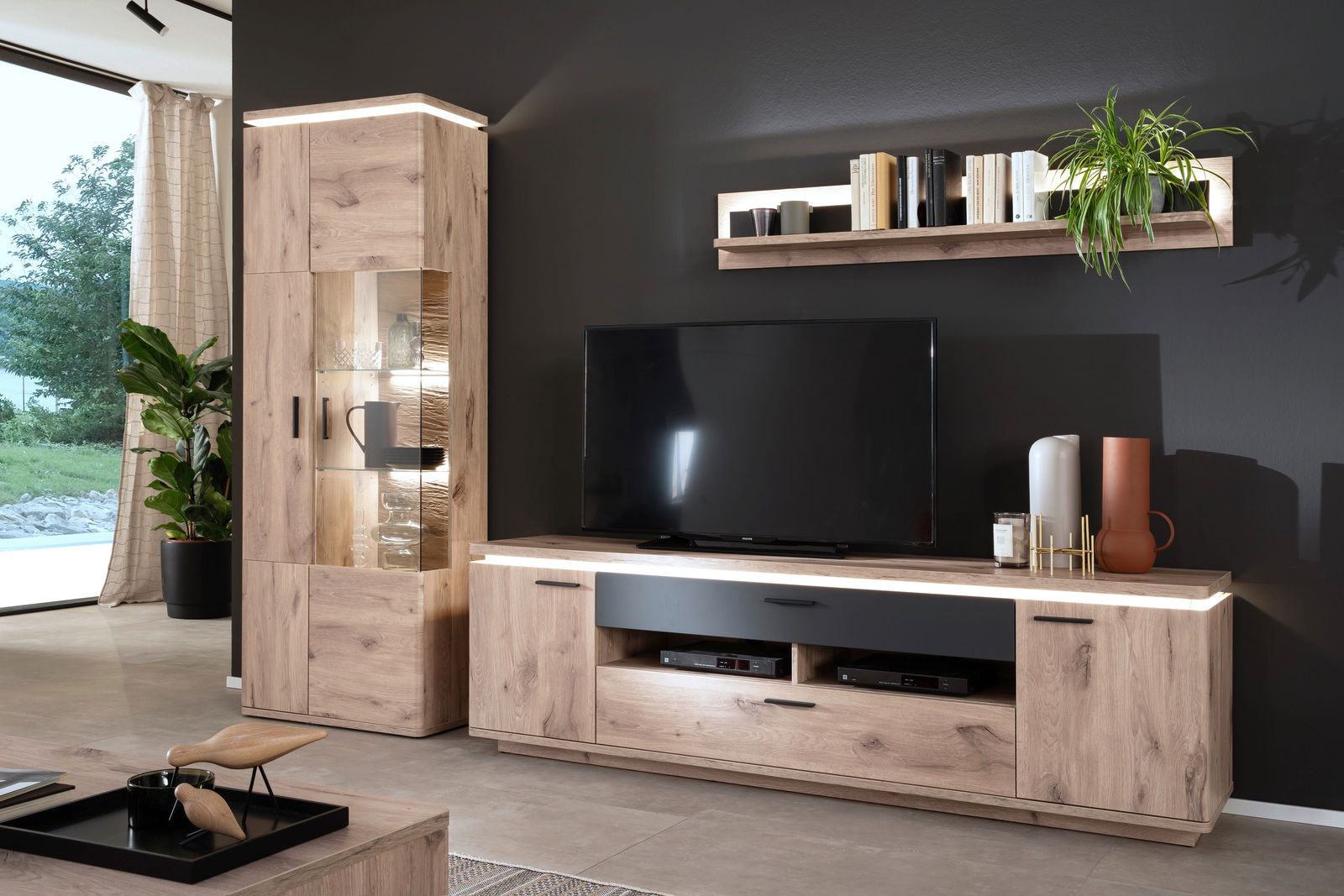 Zurbrüggen Wandkombination mit Beleuchtung MODENA Moderne Wohnzimmer-Wandkombination in Holzoptik mit LED-Beleuchtung, bestehend aus einem TV-Board, einem Highboard mit Glaselementen und einem Wandregal, seitliche Perspektive.