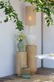 Beige Tischlampe mit zylindrischem Stoffschirm und weißem Keramikfuß auf einem Holzpodest, seitliche Perspektive im hellen Outdoor-Setting mit grünen Blättern und Deko-Vase im Vordergrund.