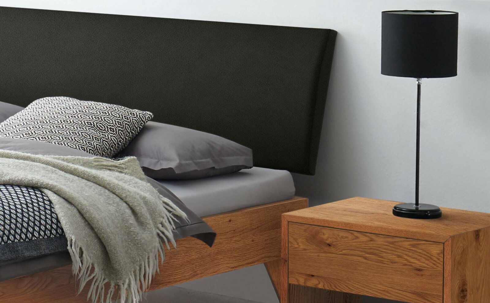 Holzbettgestell mit schwarzem Kopfteil, grauer Bettwäsche und Nachttisch mit Lampe, seitliche Perspektive
