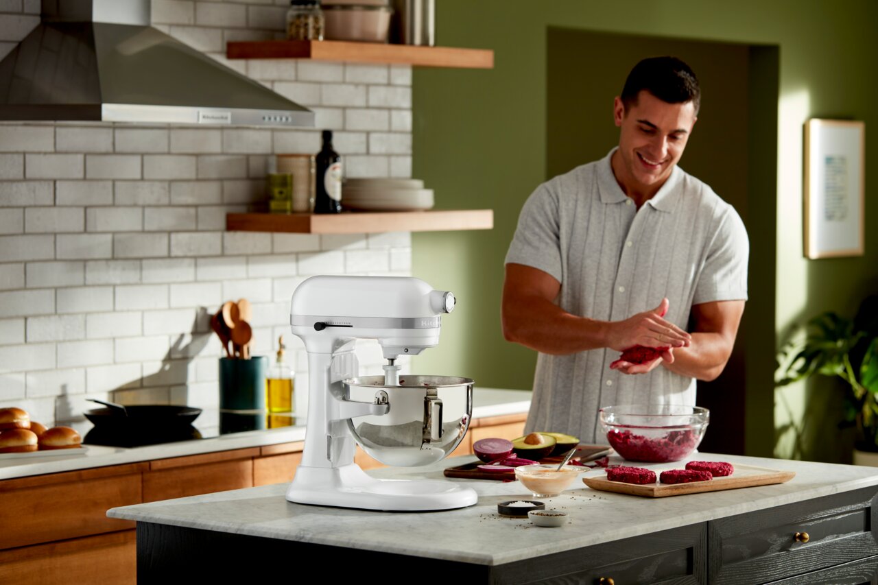 KitchenAid Küchenmaschine 5,2L HEAVY DUTY 5KSM55SXXEWH Weiße Küchenmaschine auf einer Küchenarbeitsplatte, seitliche Perspektive, mit einem Mann im Hintergrund, der Burger-Patties formt.