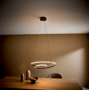 Moderne LED-Pendelleuchte mit zwei schwebenden, ringförmigen Lichtkreisen über einem Holztisch im Esszimmer, aufgenommen aus leicht niedriger Perspektive nach oben (Untersicht).