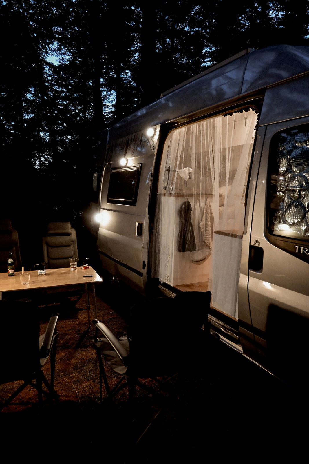Seitliche Außenansicht eines grauen Campervans am Abend im Wald, mit geöffneter Schiebetür und Vorhang, eingeschalteter Außenbeleuchtung sowie Campingtisch und Stühlen im Vordergrund (Perspektive: seitlich schräg von vorn).