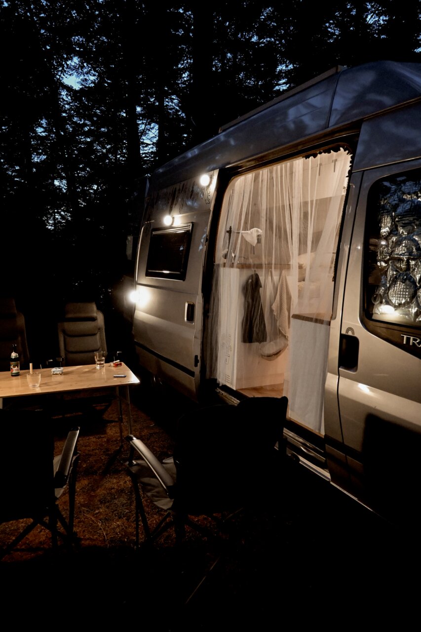 Seitliche Außenansicht eines grauen Campervans am Abend im Wald, mit geöffneter Schiebetür und Vorhang, eingeschalteter Außenbeleuchtung sowie Campingtisch und Stühlen im Vordergrund (Perspektive: seitlich schräg von vorn).