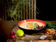 Leicht von oben fotografierte Bowl mit Reis, Lachs, Rotkohl und frischen Sprossen auf einem Holzbrett, umgeben von Limette, Radieschen und buntem Gemüse.