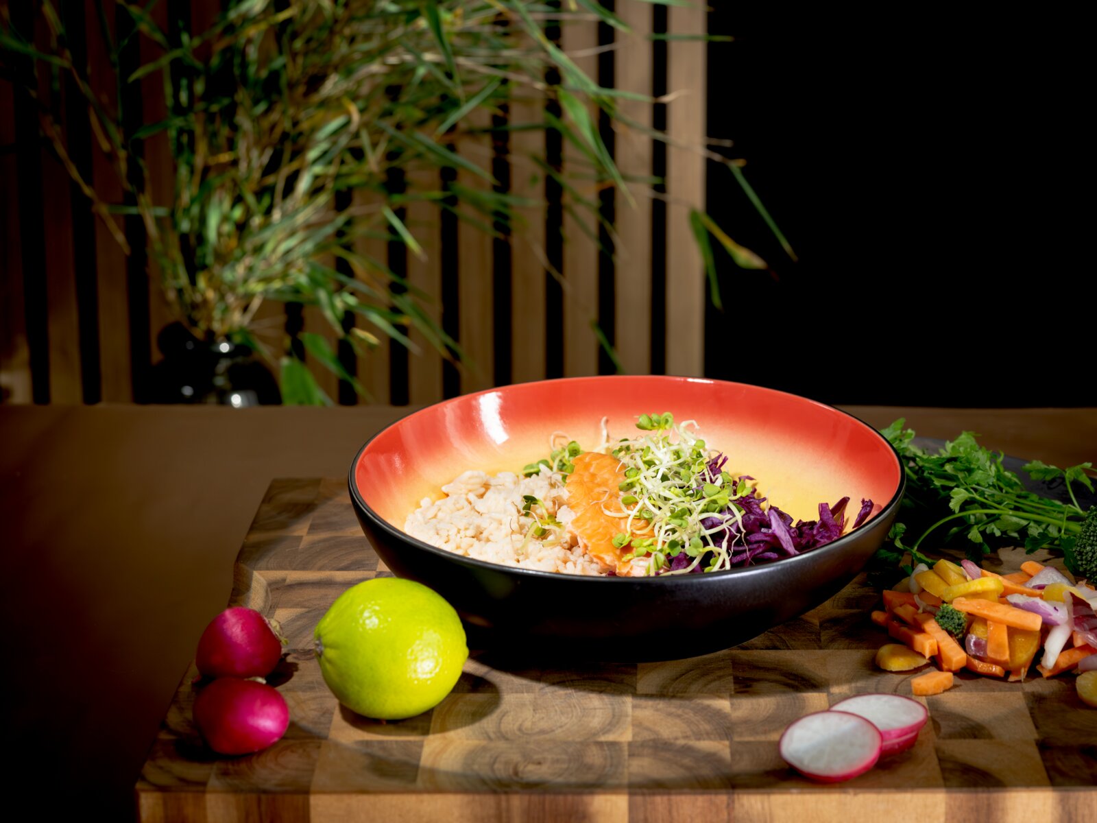 Leicht von oben fotografierte Bowl mit Reis, Lachs, Rotkohl und frischen Sprossen auf einem Holzbrett, umgeben von Limette, Radieschen und buntem Gemüse.