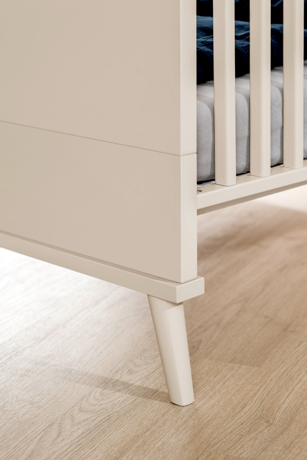 Detailaufnahme eines weißen Babybetts/Kinderbetts mit Gitterstäben und schrägem Holzfuß, seitliche Nahperspektive von unten
