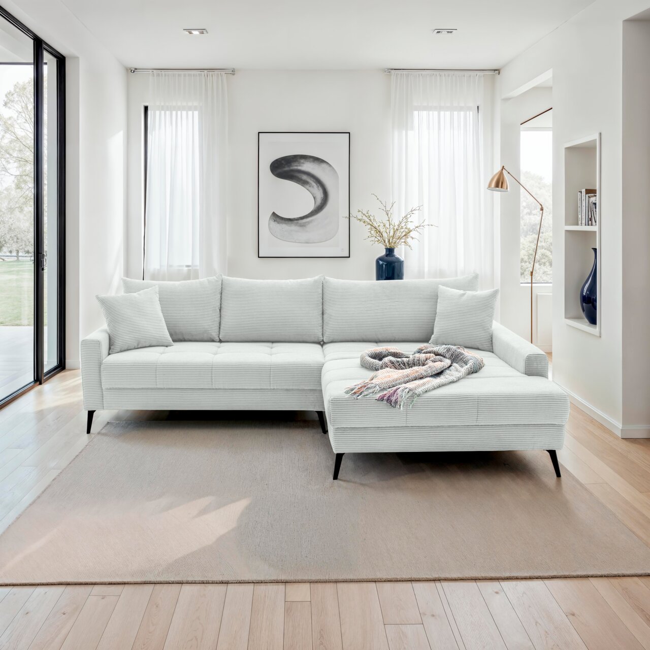 Hom´in Ecksofa SOFIA Helle, moderne Rundecke in einem stilvollen Wohnzimmer, von vorne betrachtet. Die Couch ist mit einem hellen Stoff bezogen und hat eine L-Form mit einer rechten Ecke. Ein dekorativer Überwurf liegt auf der rechten Seite der Couch.