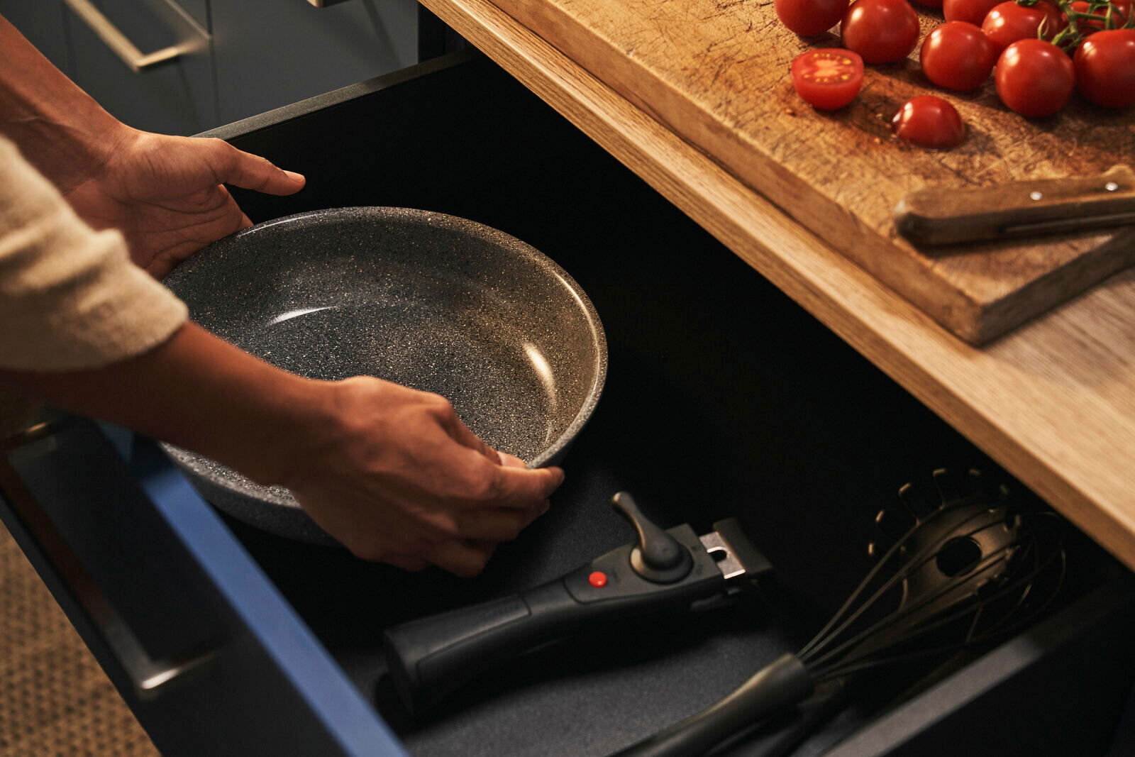 BALLARINI Pfannenset 2-tlg. Nahaufnahme einer Person, die eine graue Pfanne aus einer Küchenschublade nimmt, mit einem Schneebesen und einem abnehmbaren Griff daneben.