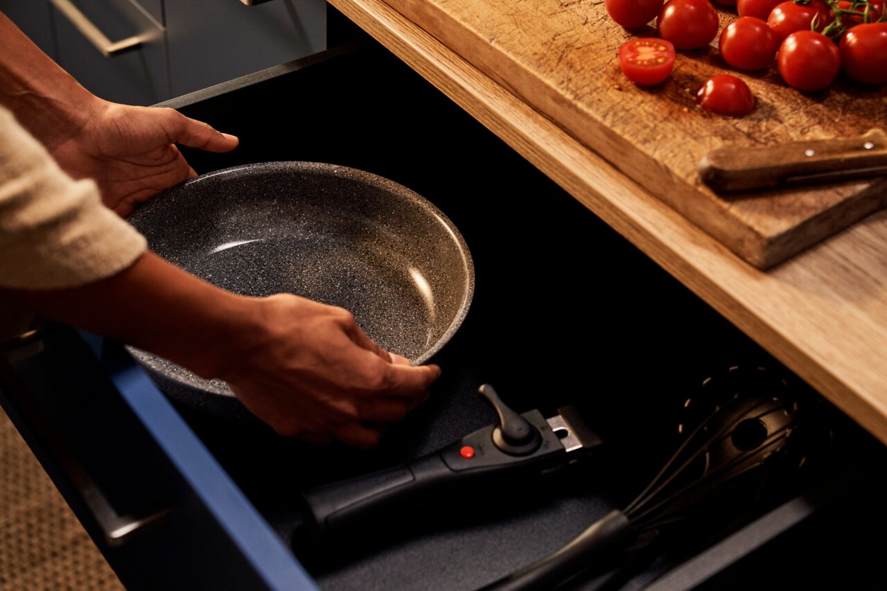 Nahaufnahme einer Person, die eine graue Pfanne aus einer Küchenschublade nimmt, mit einem Schneebesen und einem abnehmbaren Griff daneben.