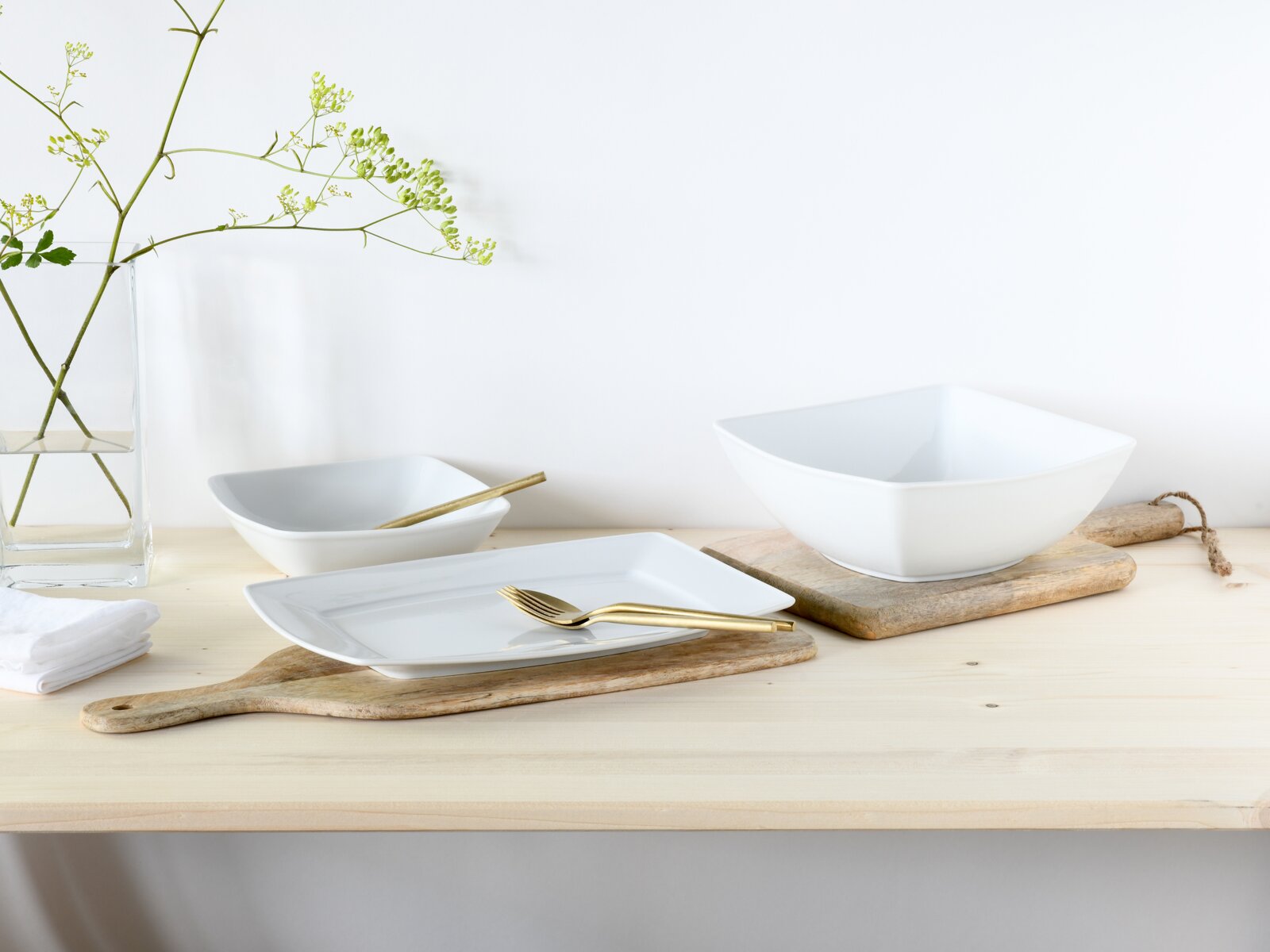 Creatable Servierset 3-tlg VICTORIA Minimalistisches weißes Geschirrset mit quadratischem Teller und Schalen auf rustikalen Holzbrettchen, goldfarbenem Besteck und Glasvase mit Zweigen auf hellem Holztisch vor weißer Wand, fotografiert aus leicht erhöhter Frontperspektive.