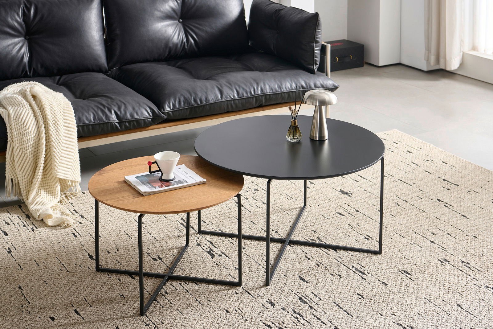 Zwei runde Couchtische im Set, einer mit schwarzer Tischplatte und der andere mit Holzoptik, auf einem hellen Teppich vor einem schwarzen Ledersofa. Perspektive von schräg oben.