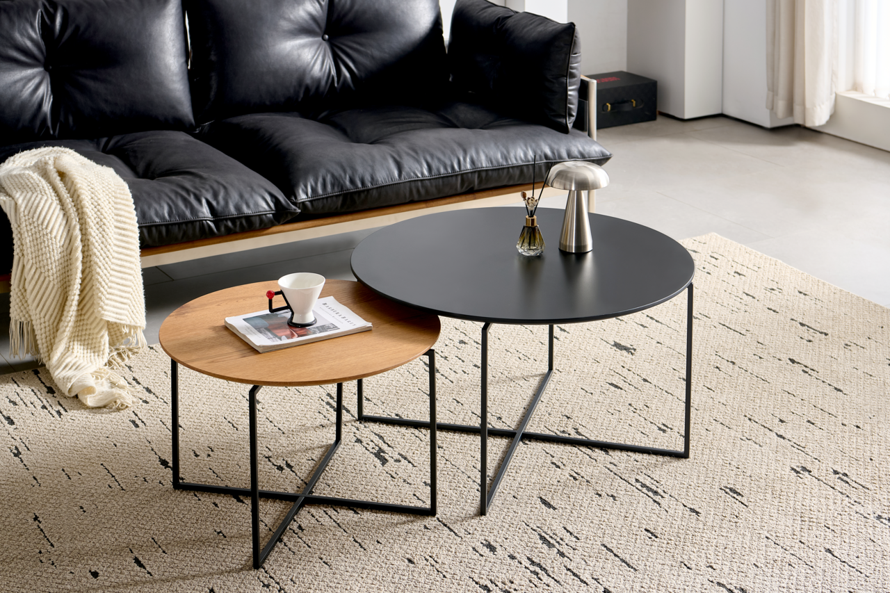 Z2 Couchtisch (Set) NICOLA Zwei runde Couchtische im Set, einer mit schwarzer Tischplatte und der andere mit Holzoptik, auf einem hellen Teppich vor einem schwarzen Ledersofa. Perspektive von schräg oben.