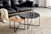 Zwei runde Couchtische im Set, einer mit schwarzer Tischplatte und der andere mit Holzoptik, auf einem hellen Teppich vor einem schwarzen Ledersofa. Perspektive von schräg oben.