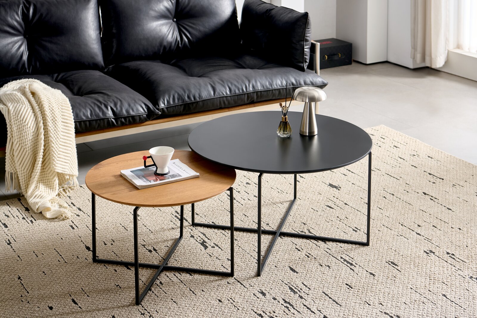 Zwei runde Couchtische im Set, einer mit schwarzer Tischplatte und der andere mit Holzoptik, auf einem hellen Teppich vor einem schwarzen Ledersofa. Perspektive von schräg oben.