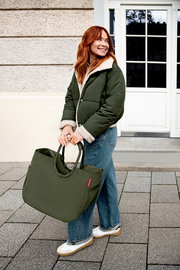 Frau trägt eine große olivgrüne Tragetasche mit runden Griffen und rotem Label, Outdoor vor einer Hausfassade; Perspektive: leicht seitliche Ganzkörperaufnahme auf Augenhöhe.