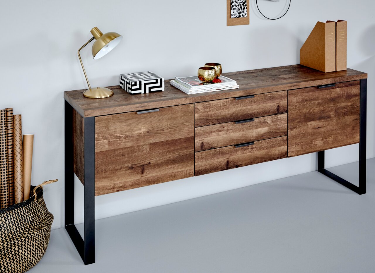 Houten dressoir met drie lades en twee deuren, op metalen poten, gefotografeerd vanuit een schuine hoek aan de voorkant. Versierd met een lamp, boeken en decoratieve voorwerpen.
