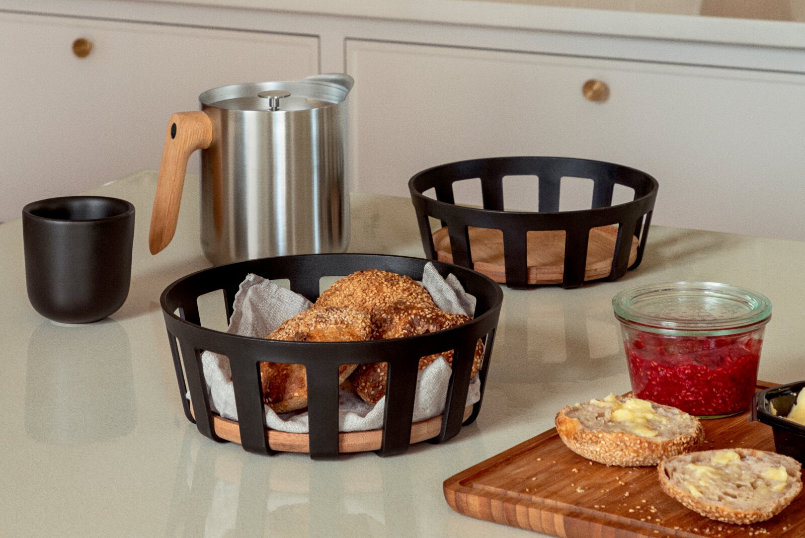 Schwarzer Brotkorb mit Holzboden und Brötchen auf einem Tisch, daneben Edelstahl-Kanne mit Holzgriff, Becher, weiterer Korb und Marmeladenglas Perspektive: leicht erhöhte Frontansicht