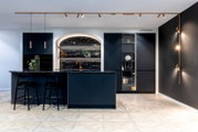 Moderne Designküche in Dunkelblau mit geriffelter Front, Kücheninsel und zwei Barhockern, schwarzer Naturstein-Arbeitsplatte, goldener Armatur und integrierter Beleuchtung, frontal aus leicht seitlicher Perspektive aufgenommen