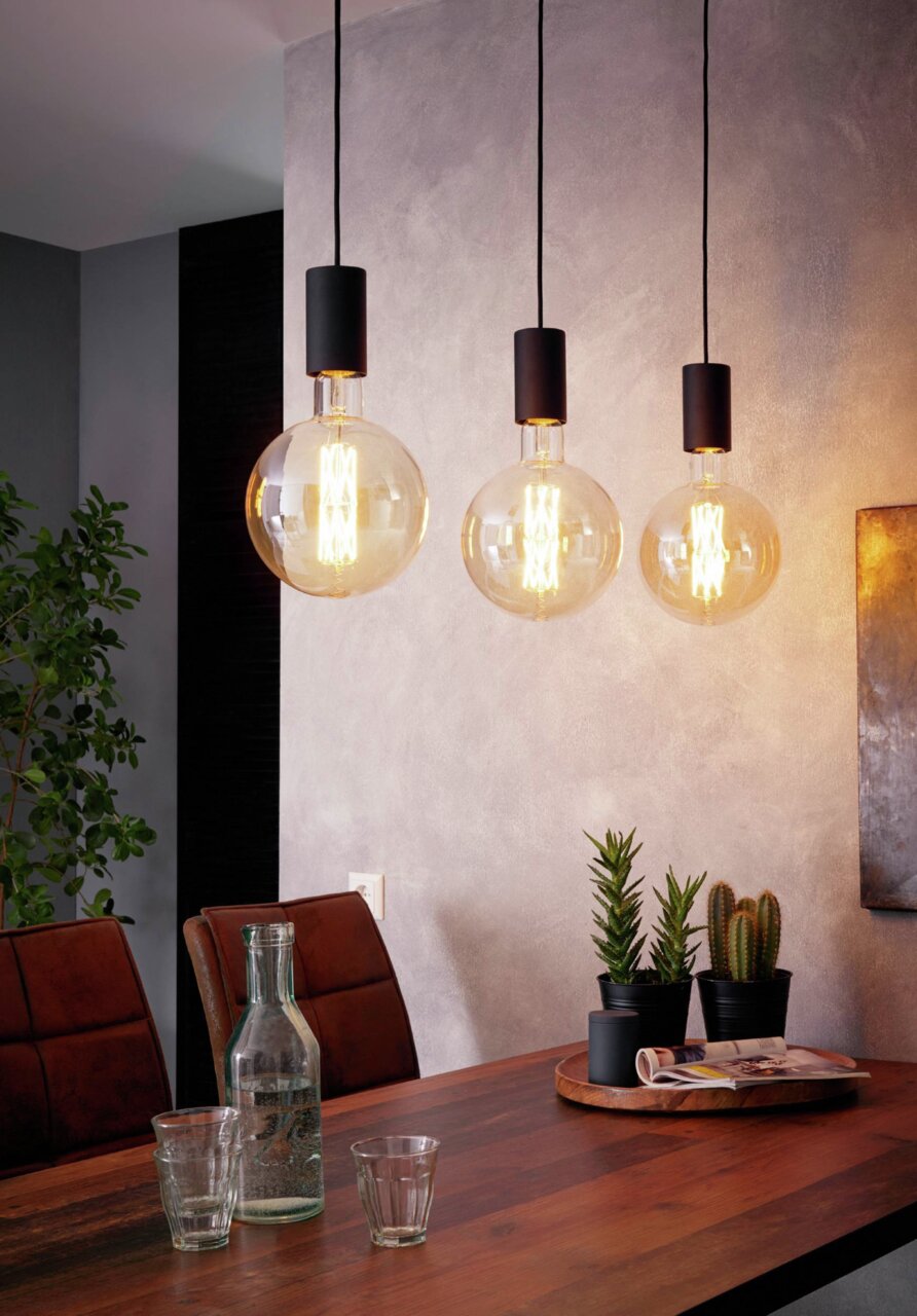 Boven een eettafel hangen drie moderne hanglampen met grote, ronde lampen. Het perspectief toont de lampen vanaf de zijkant in een stijlvol ingerichte ruimte.