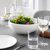 Elegante Tischszene mit einem großen weißen Salatschüssel im Vordergrund, umgeben von Weingläsern und Tellern, aus seitlicher Perspektive aufgenommen.